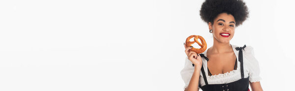 happy african american oktoberfest waitress in dirndl posing with tasty pretzel on white, banner