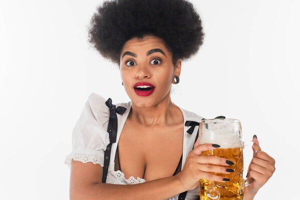 surprised african american waitress in traditional bavarian dirndl holding beer mug on white