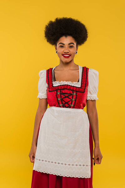 pleased african american waitress in authentic bavarian costume on yellow, oktoberfest concept