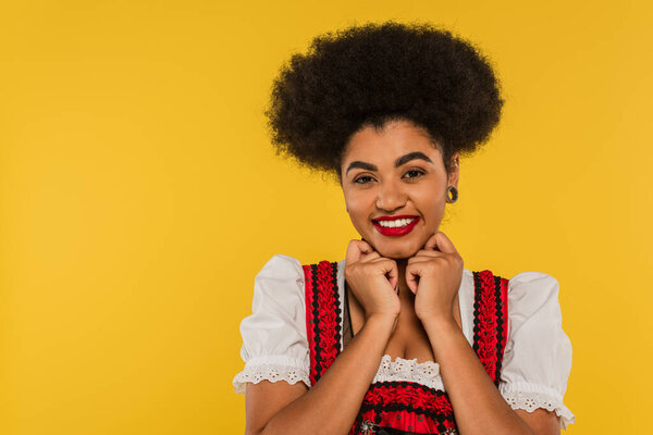 overjoyed african american oktoberfest waitress in traditional dirndl looking at camera on yellow