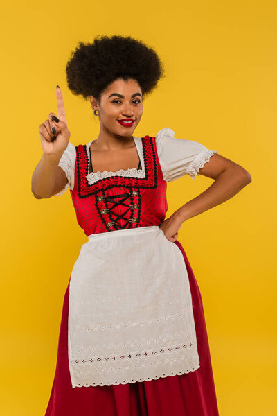 smiling african american bavarian waitress with hand on hip showing idea gesture on yellow