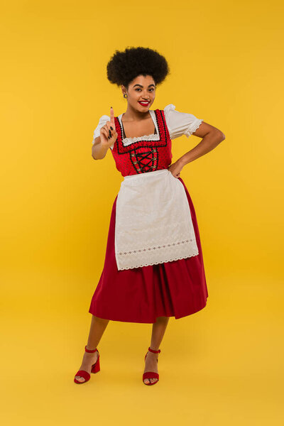 happy african american oktoberfest waitress with hand on hip showing idea sign on yellow