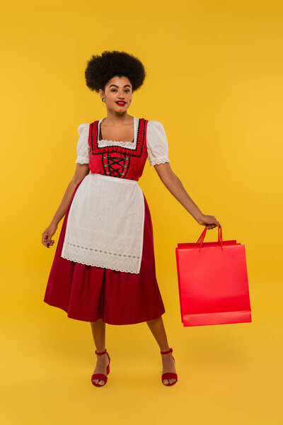 pleased african american bavarian waitress in oktoberfest outfit holding shopping bags on yellow