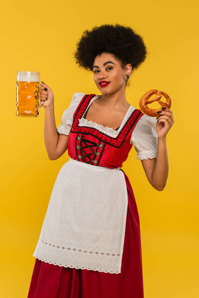 african american oktoberfest waitress with beer mug and tasty pretzel smiling at camera on yellow