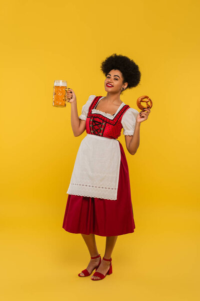 joyful african american waitress in bavarian costume with beer mug and delicious pretzel on yellow