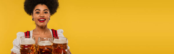excited african american bavarian waitress smiling at camera near beer mugs on yellow, banner