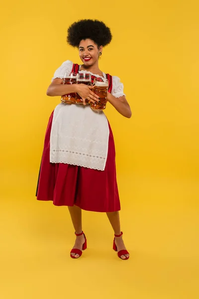 joyful african american oktoberfest waitress with full beer mags looking at camera on yellow