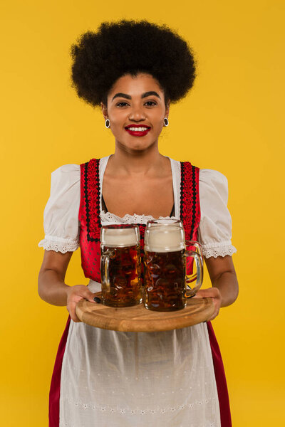 happy african american waitress in oktoberfest dress serving bavarian beer in mugs on tray on yellow