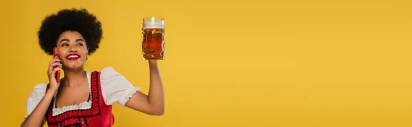 happy african american bavarian waitress with beer mug talking on smartphone on yellow, banner