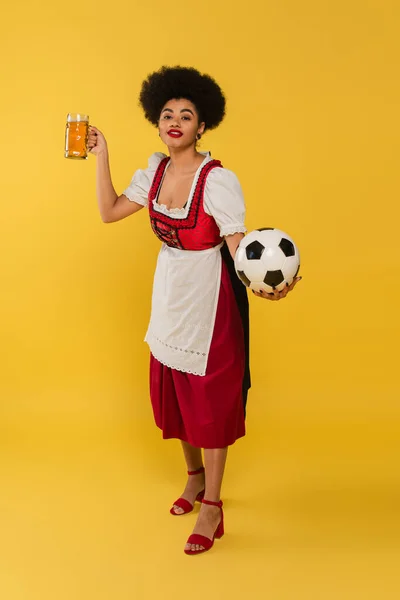 stock image pleased african american waitress in bavarian costume with beer mug and soccer ball on yellow