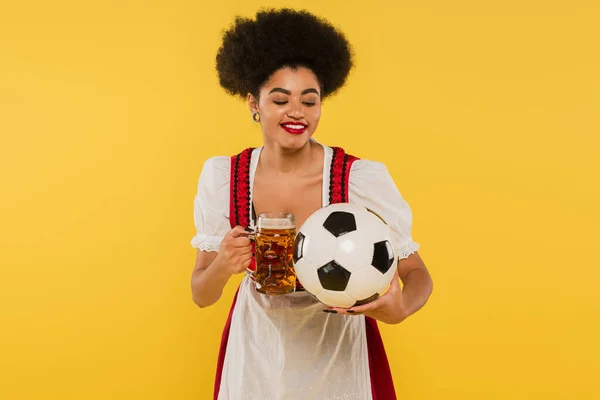 stock image joyful african american oktoberfest waitress in dirndl clinking beer mug with soccer ball on yellow