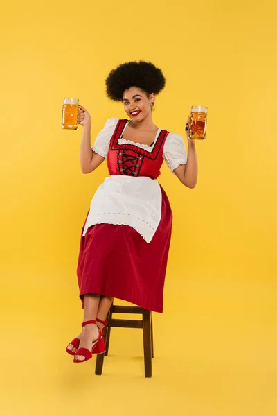 excited african american bavarian waitress in dirndl sitting on chair with beer mugs on yellow