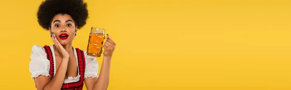 stock image amazed african american oktoberfest waitress with beer mug looking at camera on yellow, banner