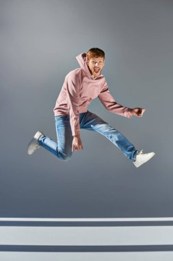 cheerful young man in casual clothing jumping and playing invisible guitar on grey backdrop clipart