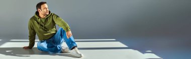 young indian man in trendy khaki hoodie sitting on floor looking away on grey backdrop, banner clipart