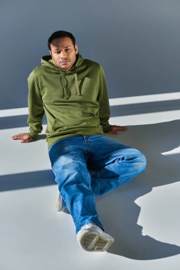 young indian man in casual urban attire slightly smiling sitting on floor and looking at camera clipart