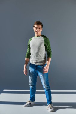 Uzun kollu ve kot pantolonlu genç ve neşeli bir adam gri arka planda kameraya bakıyor.