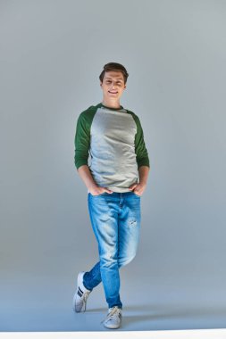 Uzun kollu ve kot pantolonlu genç adam kameraya gülümsüyor elleri ceplerinde poz veriyor.