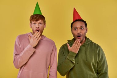 astonished multicultural men in everyday street wear in birthday hats covering mouths with hands clipart