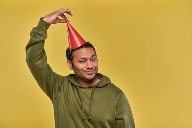 Sokak kıyafetli gülümseyen Hintli adam kurnaz bakışlarla sarı arka planda doğum günü şapkasına dokunuyor.