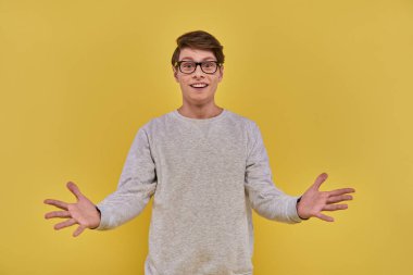 cheerful surprised man in casual attire and glasses raising hands with open palms on yellow backdrop clipart