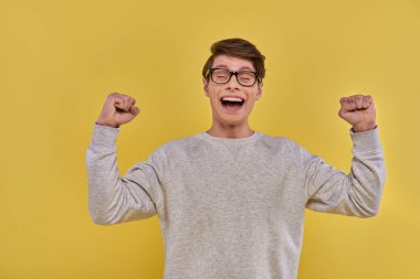 emotional happy man in casual street outfit raising fists up and closing his eyes on yellow backdrop clipart