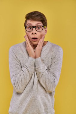 shocked young man in casual attire with glasses touching his cheeks with hands on yellow backdrop clipart