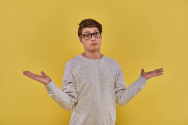 confused clueless young man in sweatshirt and glasses showing helpless gesture on yellow backdrop clipart