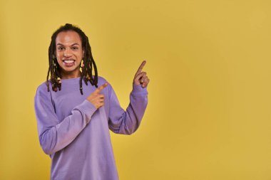 cheerful african american man with dreadlocks pierced lip pointing on right side on yellow backdrop clipart