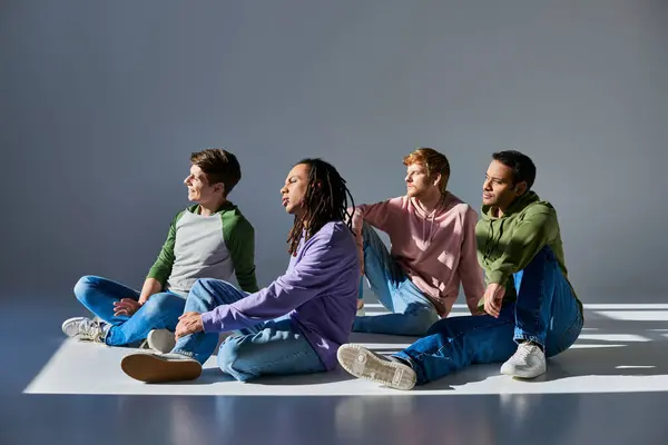 four young men in casual outfits sitting on floor on grey background and looking away, diversity