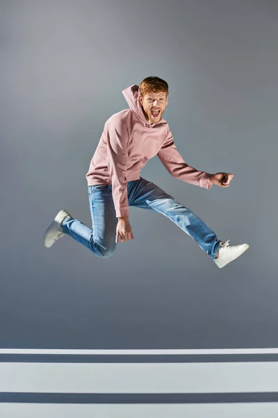stock image cheerful young man in casual clothing jumping and playing invisible guitar on grey backdrop