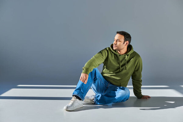 indian man in casual street wear sitting on floor and looking away on grey background