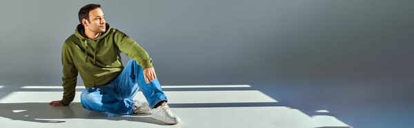 young indian man in trendy khaki hoodie sitting on floor looking away on grey backdrop, banner