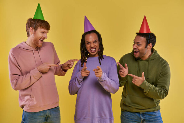 three multicultural friends in birthday hats celebrating and pointing fingers at camera, birthday
