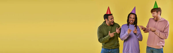 three multiethnic men in birthday hats smiling and pointing fingers at camera, birthday, banner