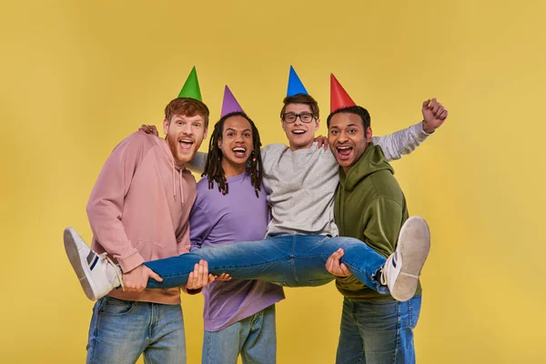 four cheerful men in birthday hats smiling at camera and holding one of them on hands, birthday