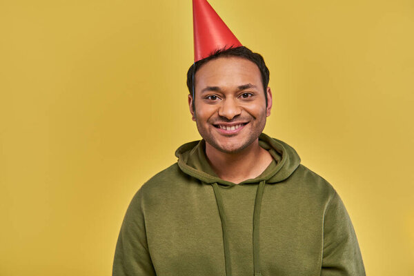 cheerful young indian man in birthday hat and casual khaki hoodie smiling at camera, birthday