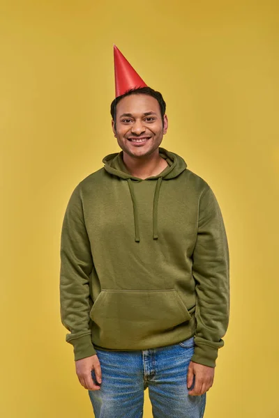 joyful indian man in casual attire and birthday hat smiling at camera on yellow backdrop, birthday