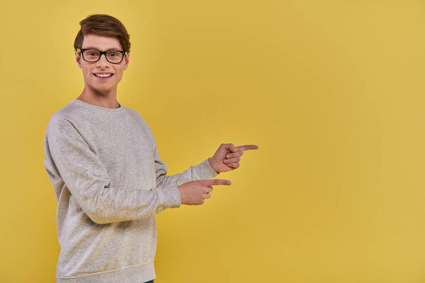 cheerful young man in casual attire pointing two fingers aside on right side on yellow backdrop