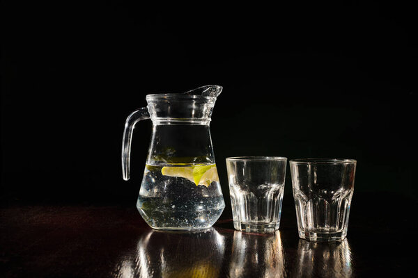 освежающий полный кувшин пресной и чистой воды с лимоном и пустыми стаканами на черном фоне