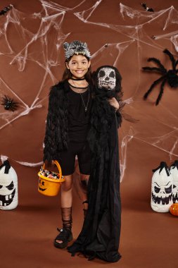 happy preteen girl in wolf mask with bucket of sweets and spooky toy, Halloween concept clipart