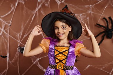 joyous girl in Halloween costume adjusting witch hat and looking at camera on brown backdrop clipart
