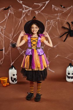 happy girl in Halloween costume adjusting witch hat and standing near cobwebs on brown backdrop clipart