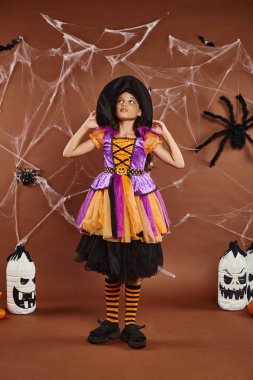 girl in witch hat and Halloween costume standing near spooky decor and cobwebs on brown backdrop clipart