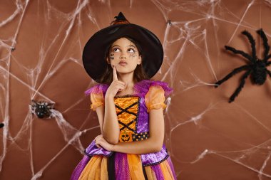 pensive girl in witch hat and Halloween costume standing near cobwebs on brown backdrop, spooky clipart