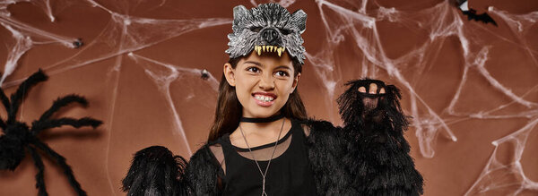 close up of preteen girl in black attire and wolf mask scaring with raised hands, Halloween concept