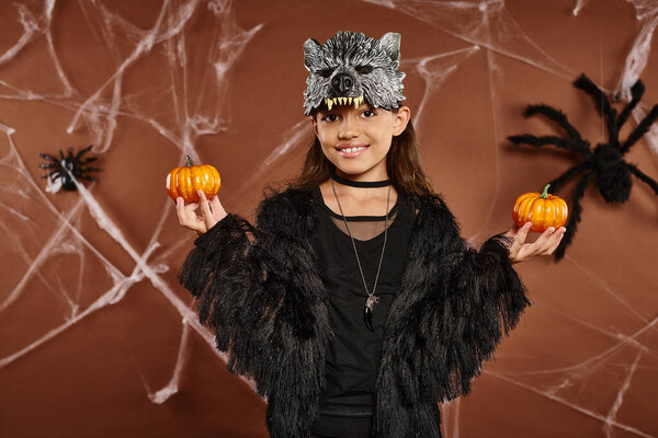 smiley girl in wolf mask holds pumpkins on brown backdrop with spiders and cobweb, Halloween concept