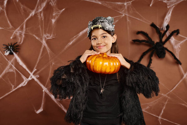 pretty preteen girl holding pumpkin under her chin on brown backdrop, Halloween concept