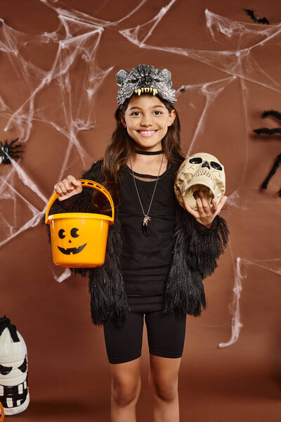 close up smiling preteen girl with skull and bucket of sweets, brown background with web, Halloween
