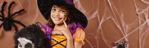 close up cheerful girl in witch hat with spooky toy touching cheek and smiling, Halloween, banner